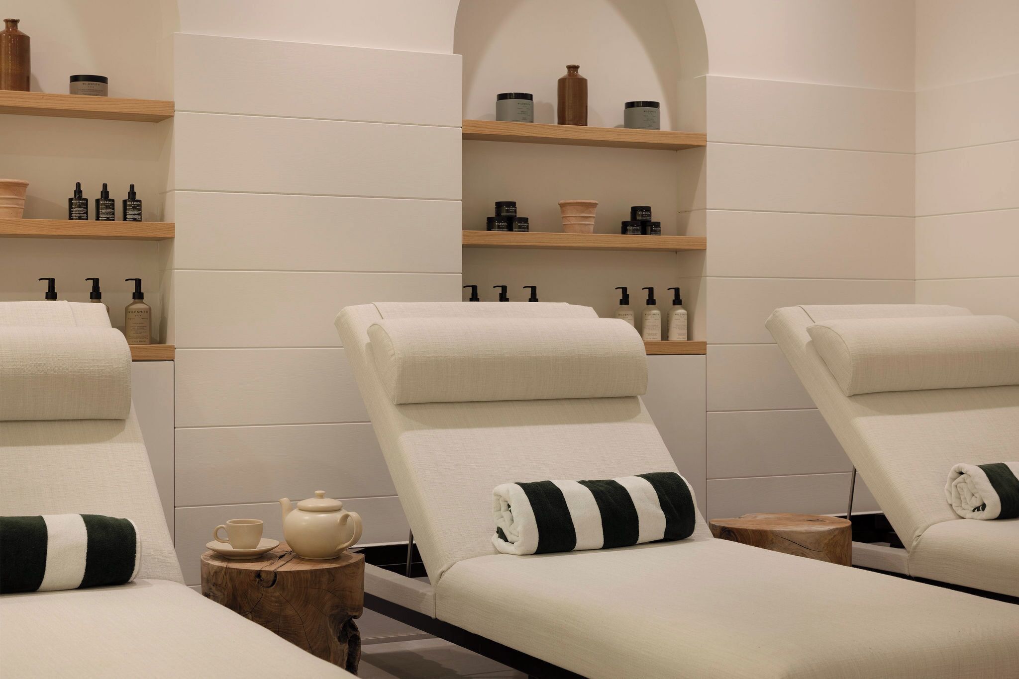 A spa room with three reclining chairs, wooden shelves with toiletries, and a small table with a teapot and cup.