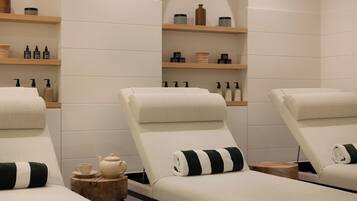 A spa room with three reclining chairs, wooden shelves with toiletries, and a small table with a teapot and cup.