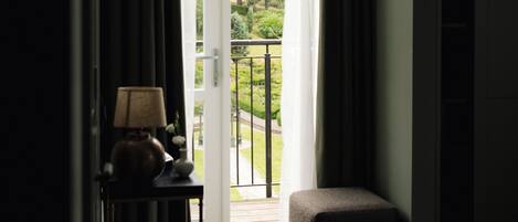 A hotel room with a view of a garden through a glass door, a bedside table with a lamp, and a small seating area.