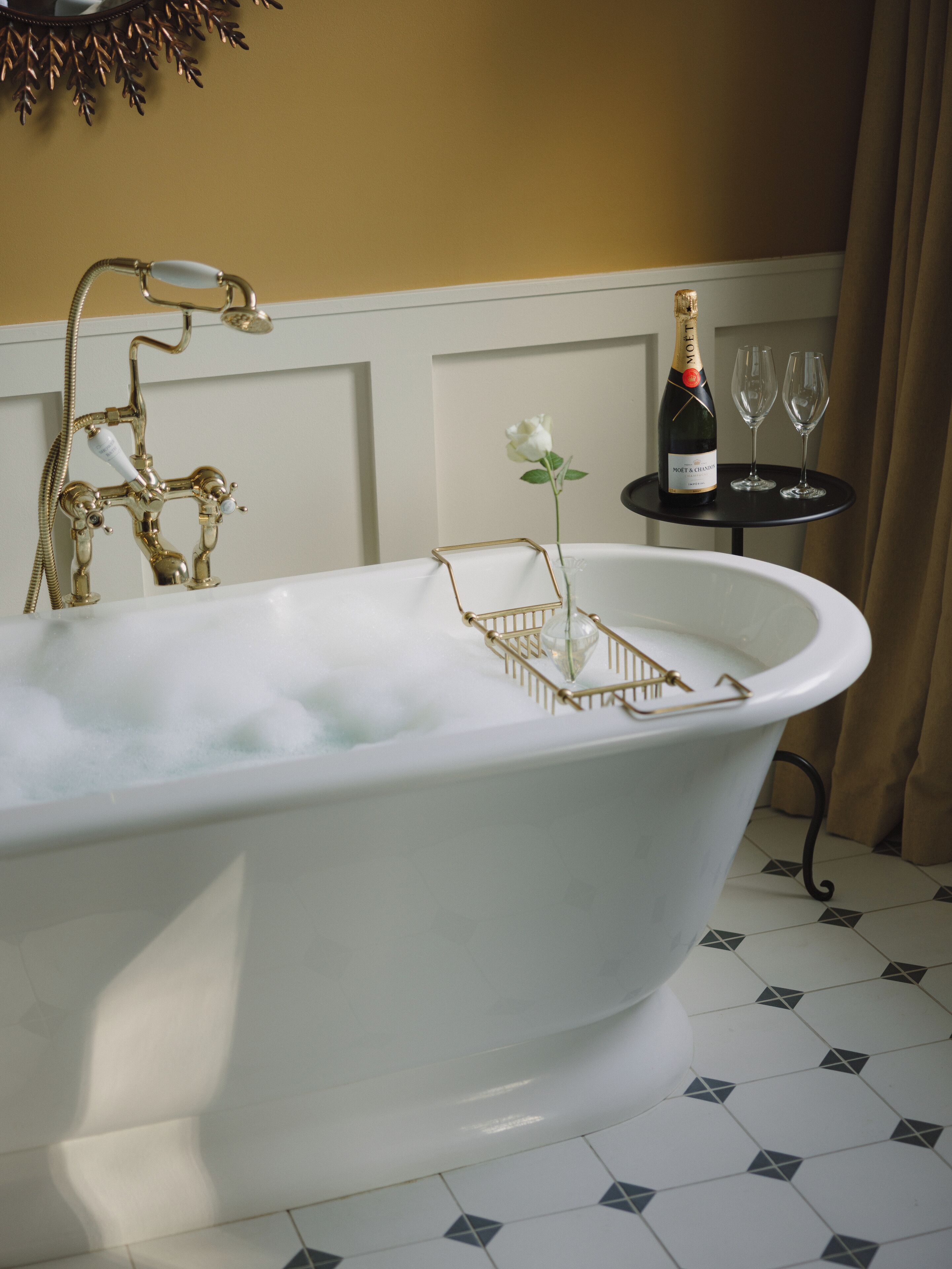 A bathtub with a gold faucet, a bottle of champagne, and two glasses on a small table beside it.