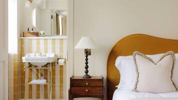 A hotel room with a bed, a bedside table with a lamp, a nightstand, a vanity with a mirror, and a towel rack.