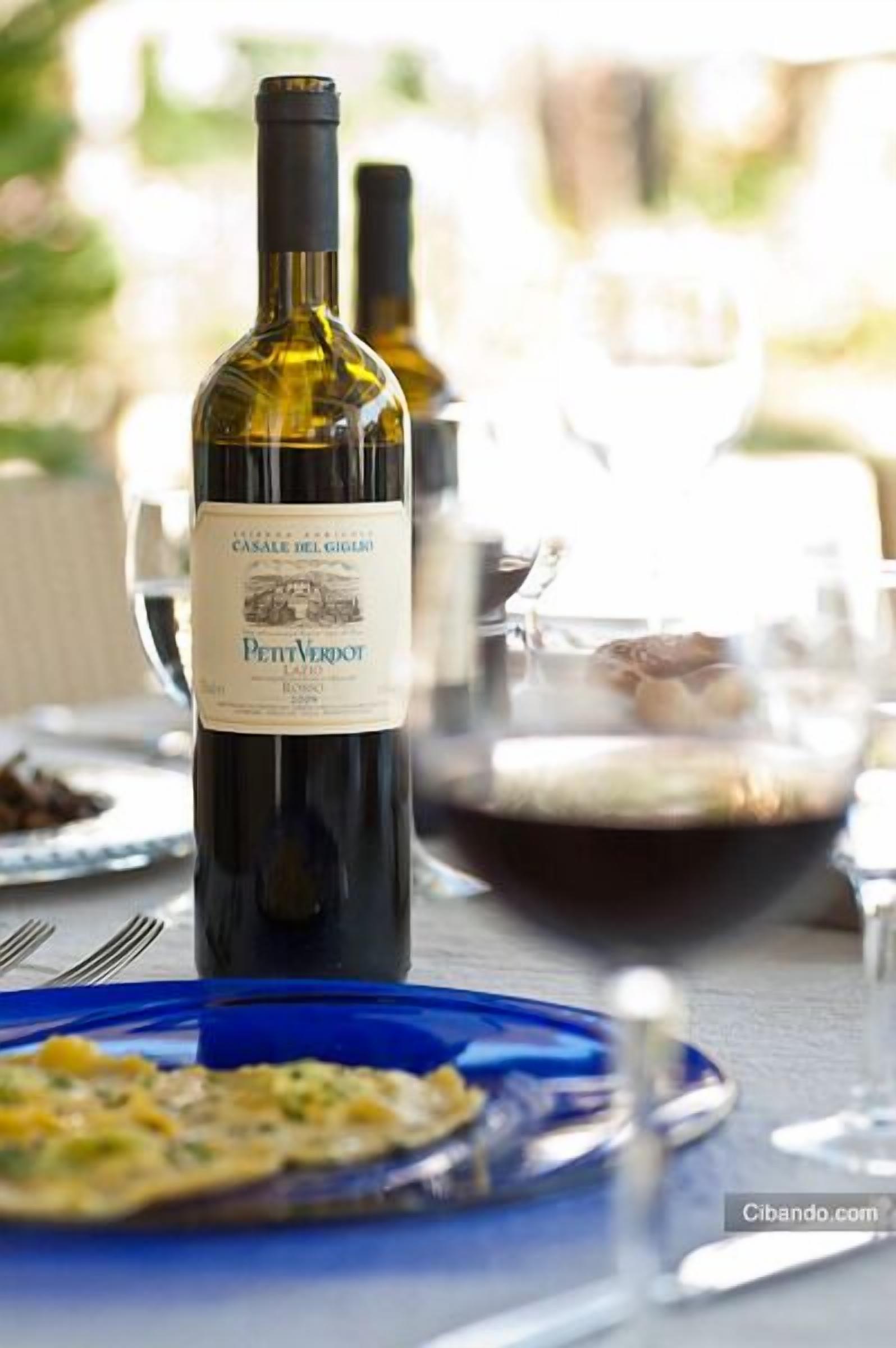 A bottle of Petit Verdot wine on a table with a blue plate of food and a glass of red wine.