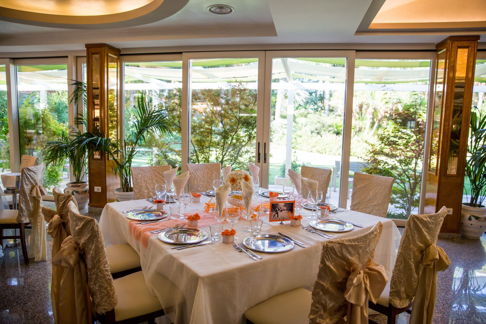 Un comedor con una mesa dispuesta para una comida, rodeado de sillas decoradas con lazos, ventanas amplias con vistas a la vegetación y plantas en macetas.