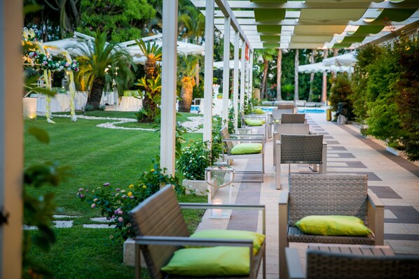 An outdoor seating area with wicker chairs and cushions, a pool, and a well-maintained garden with palm trees and flowers.