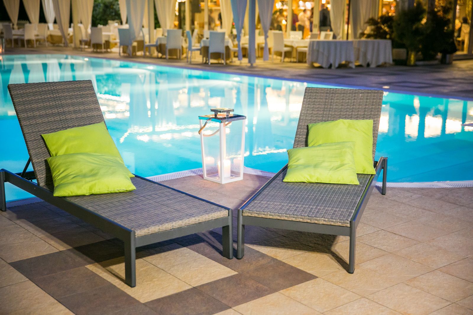 Two lounge chairs with green cushions by a poolside, a small table with candles, and a dining area in the background.