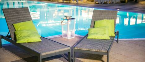 Two lounge chairs with green cushions by a poolside, a small table with candles, and a dining area in the background.