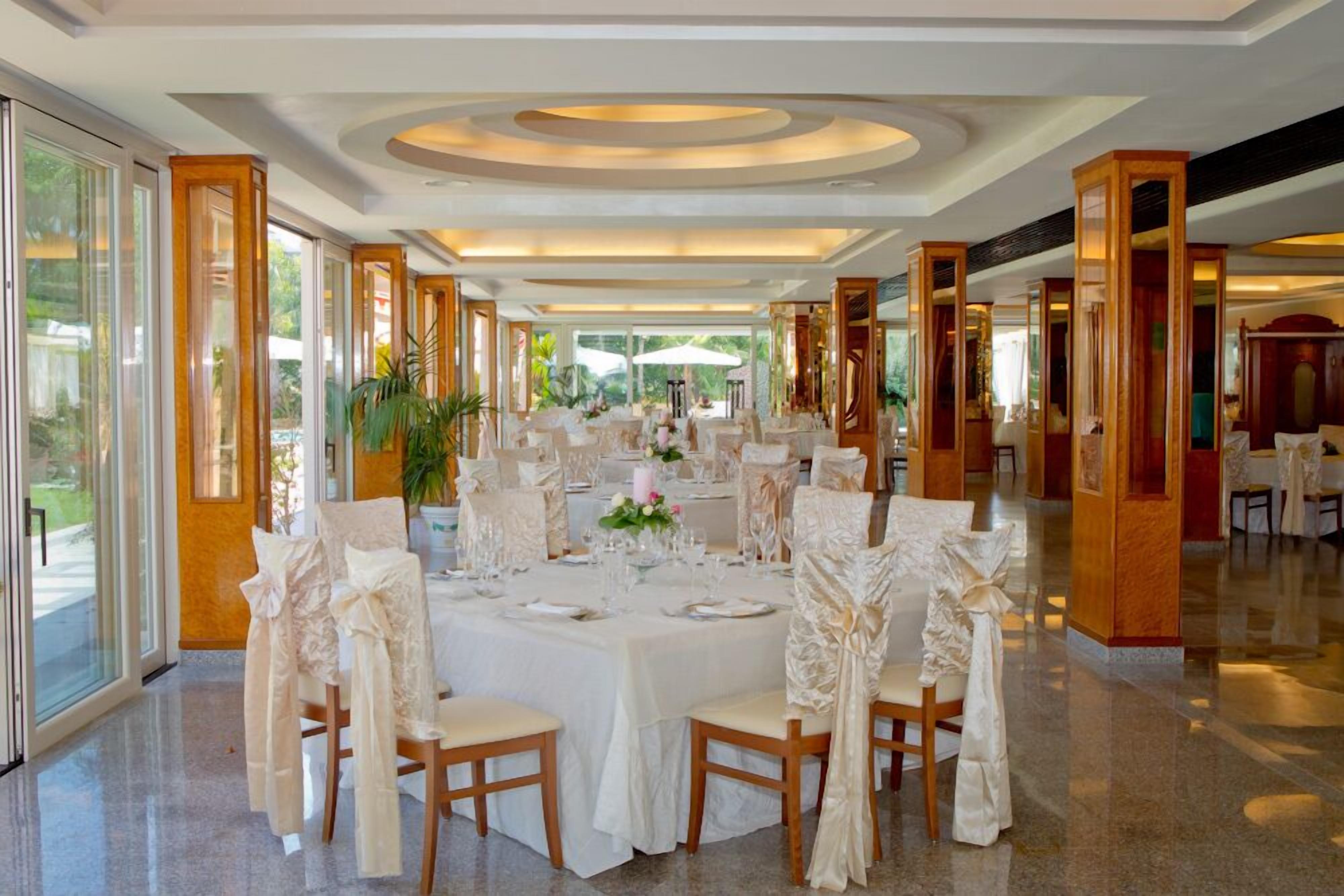 Une salle à manger spacieuse, avec des tables rondes, des nappes blanches et des chaises ornées de rubans blancs.