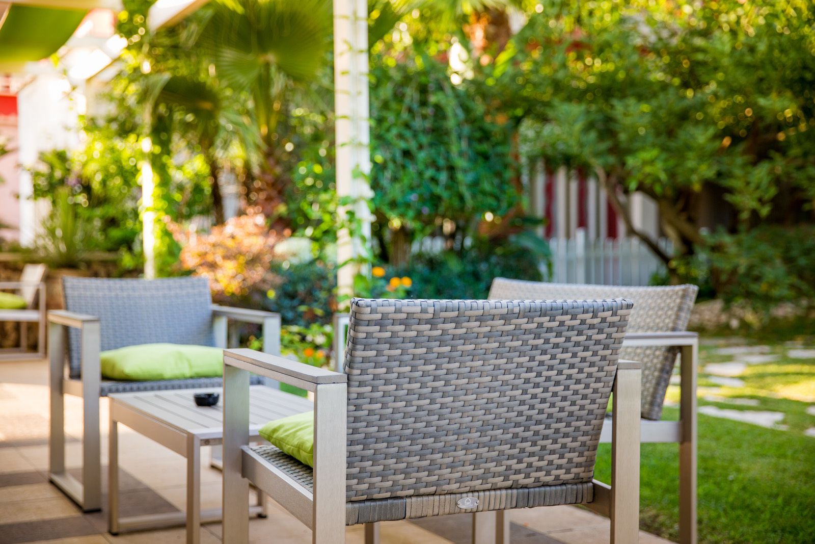 An outdoor seating area with wicker chairs and a table, surrounded by greenery and a white fence.