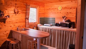 A wooden cabin kitchen with a wooden bar, stools, a sink, a microwave, and a window with plaid curtains. - Les Chalets du Trappeur avec Spa et Sauna (Wentworth-Nord)
