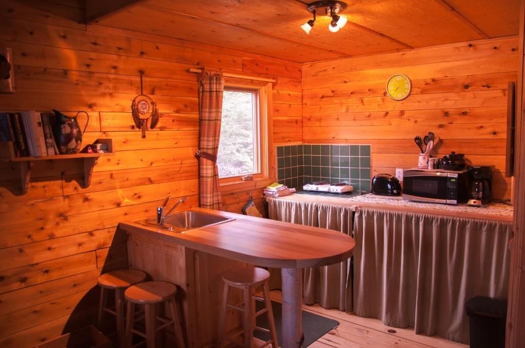 L’intérieur d’un chalet en bois comprend un coin cuisine, une table à manger avec des tabourets et une fenêtre avec des rideaux.