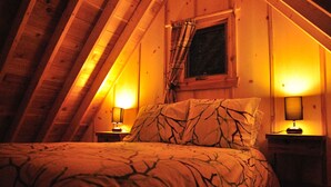 A cozy attic bedroom with a sloped ceiling, a bed with patterned bedding, two bedside lamps, and a window with curtains. - Les Chalets du Trappeur avec Spa et Sauna (Wentworth-Nord)