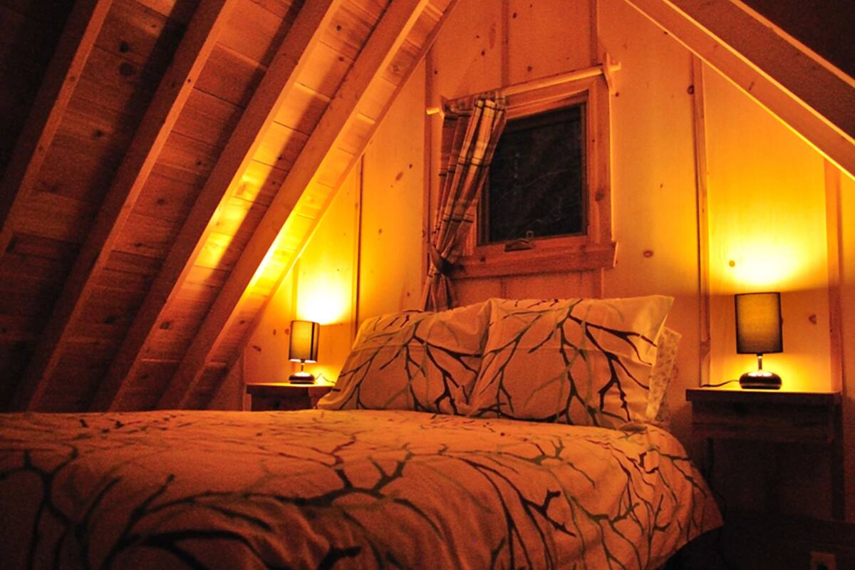 Un dormitorio acogedor en el ático, con techo inclinado, una cama con ropa de cama estampada, dos lámparas de noche y una ventana con cortinas.