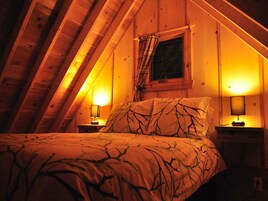 Un dormitorio acogedor en el ático, con techo inclinado, una cama con ropa de cama estampada, dos lámparas de noche y una ventana con cortinas.