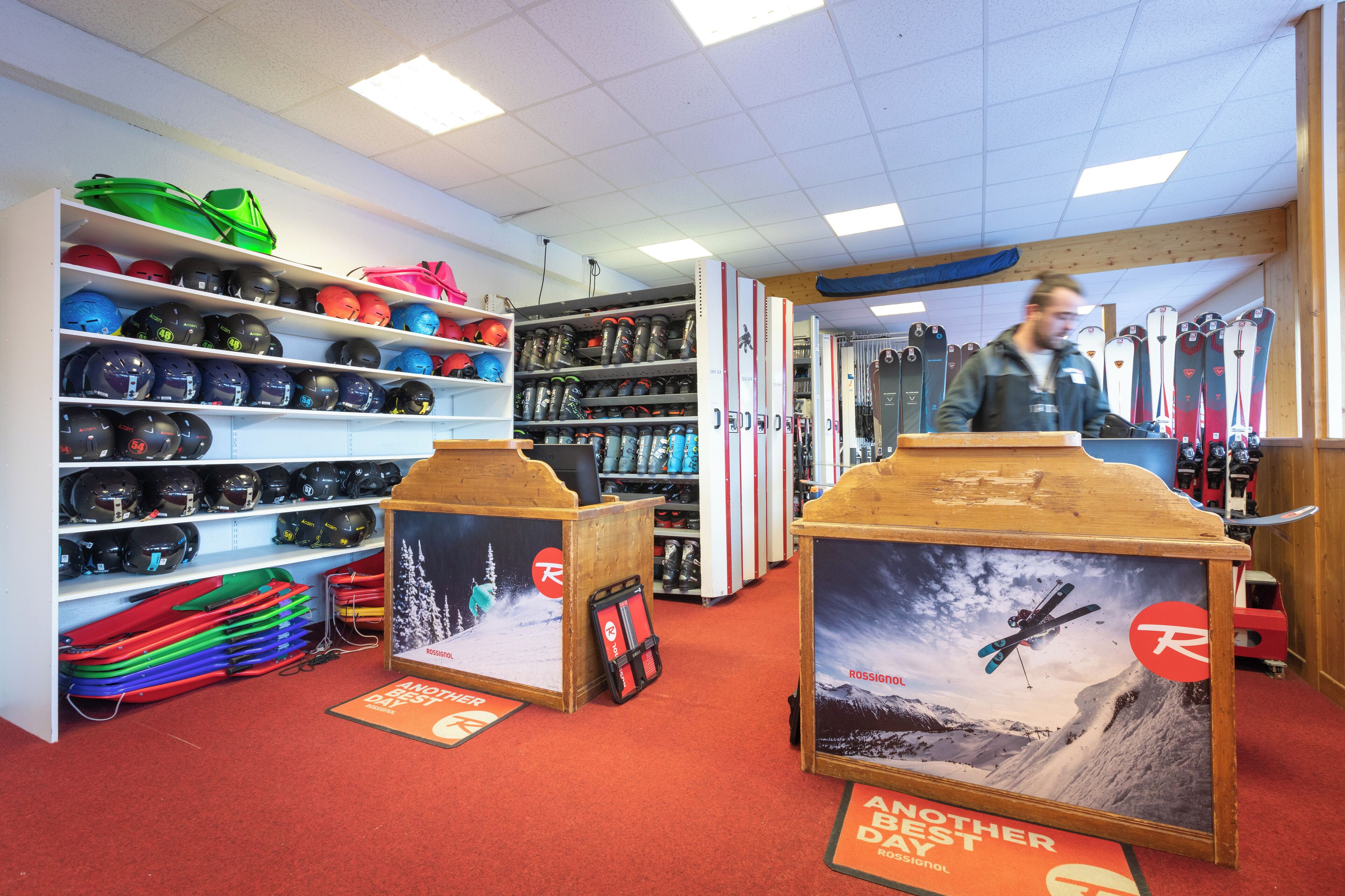A ski equipment store with shelves of helmets and goggles, a display of skis, and a poster advertising a Rossignol ski brand.