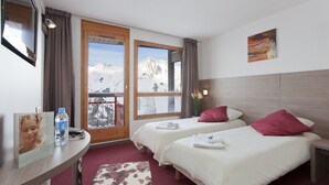 Ein Hotelzimmer mit zwei Betten, einem Holz-Kopfstück, einem Fenster mit Blick auf die Berge, einem kleinen Tisch mit einer Wasserflasche und einem gerahmten Bild an der Wand.