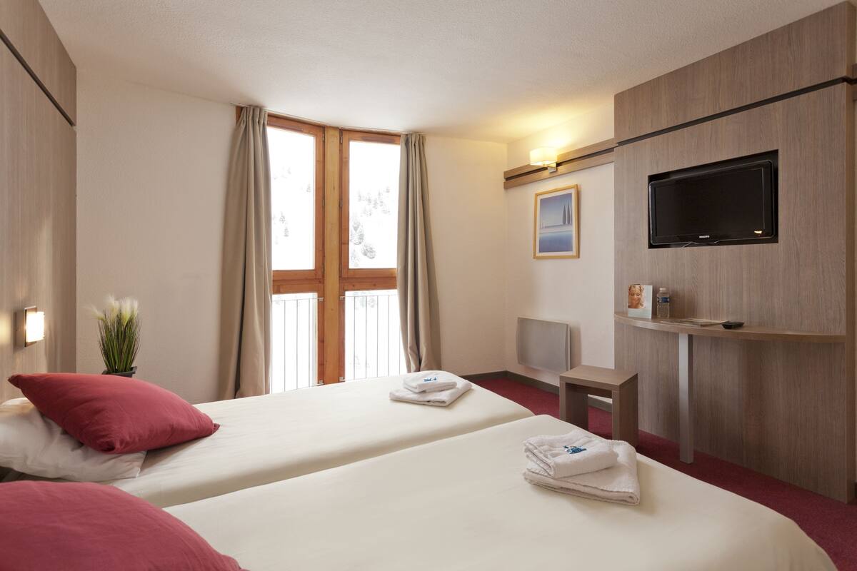 Ein Hotelzimmer mit zwei Betten, einem Fernseher, einem Schreibtisch und einem fensterbepflanzten Fenster.