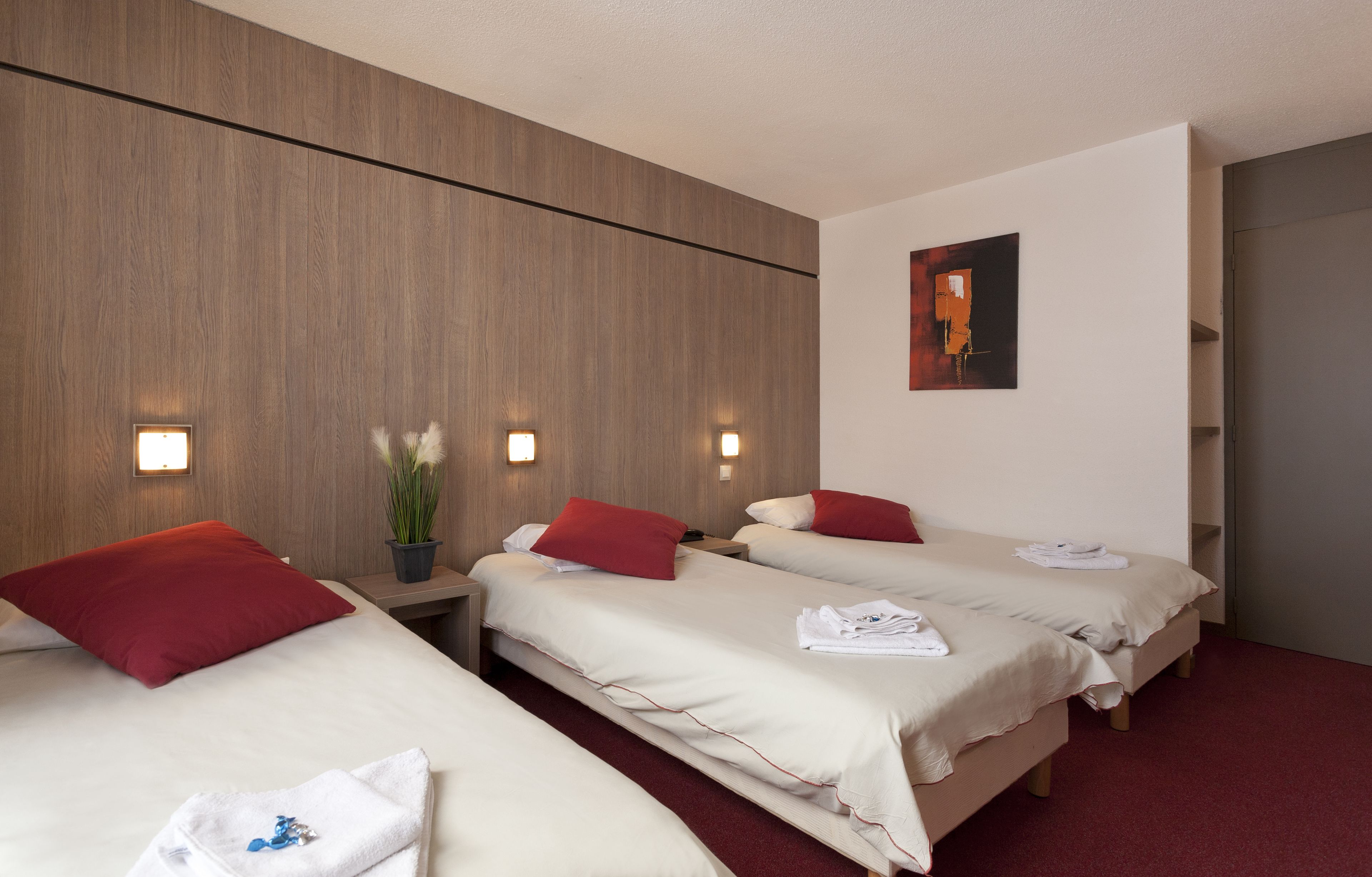 A hotel room with three single beds, each with red pillows and white linens. There is a wooden panel wall, a small potted plant, and a painting on the wall.