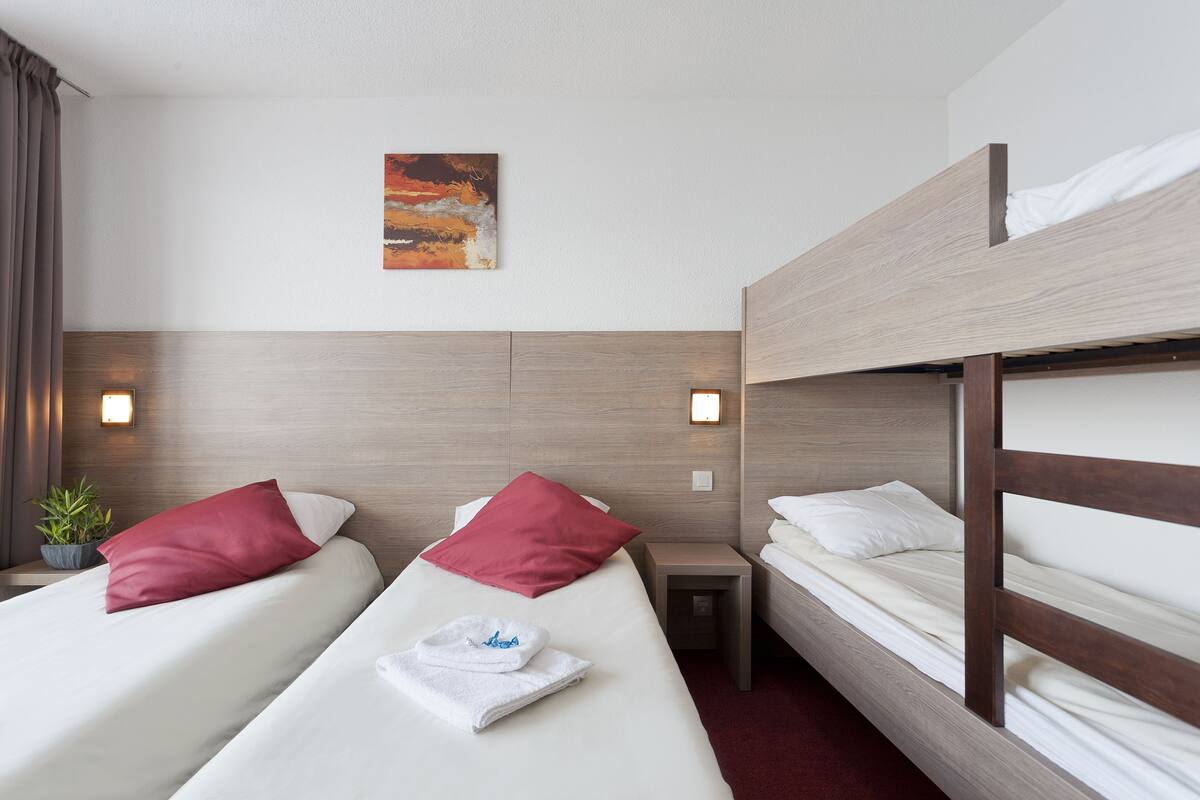 Ein Hotelzimmer mit zwei Einzelbetten, einem Etagenbett, einem kleinen Nachttisch mit Lampe, einem Bild an der Wand und einer Zimmerpflanze.