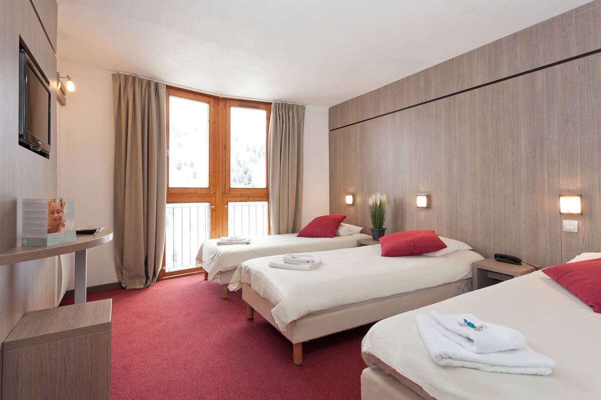 Ein Hotelzimmer mit drei Betten, einem Schreibtisch und einem Fenster mit Vorhängen.
