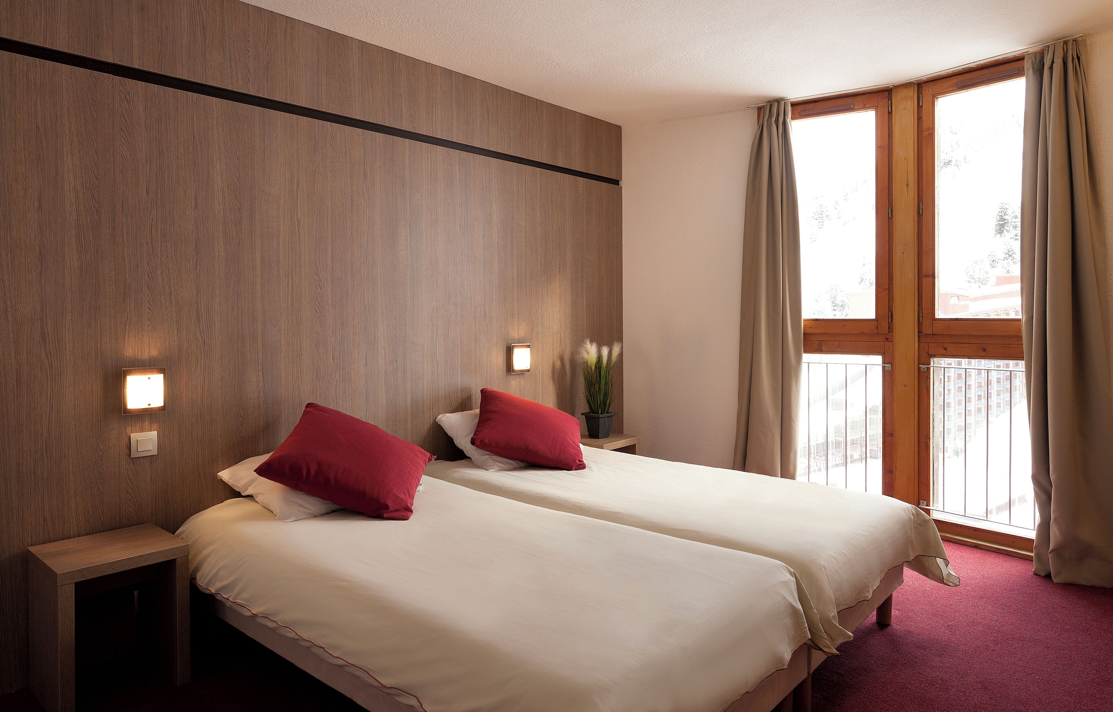 Ein Hotelzimmer mit zwei Betten, einem Nachttisch, einer Wandlampe, einem Fenster mit Vorhängen und Blick ins Freie.