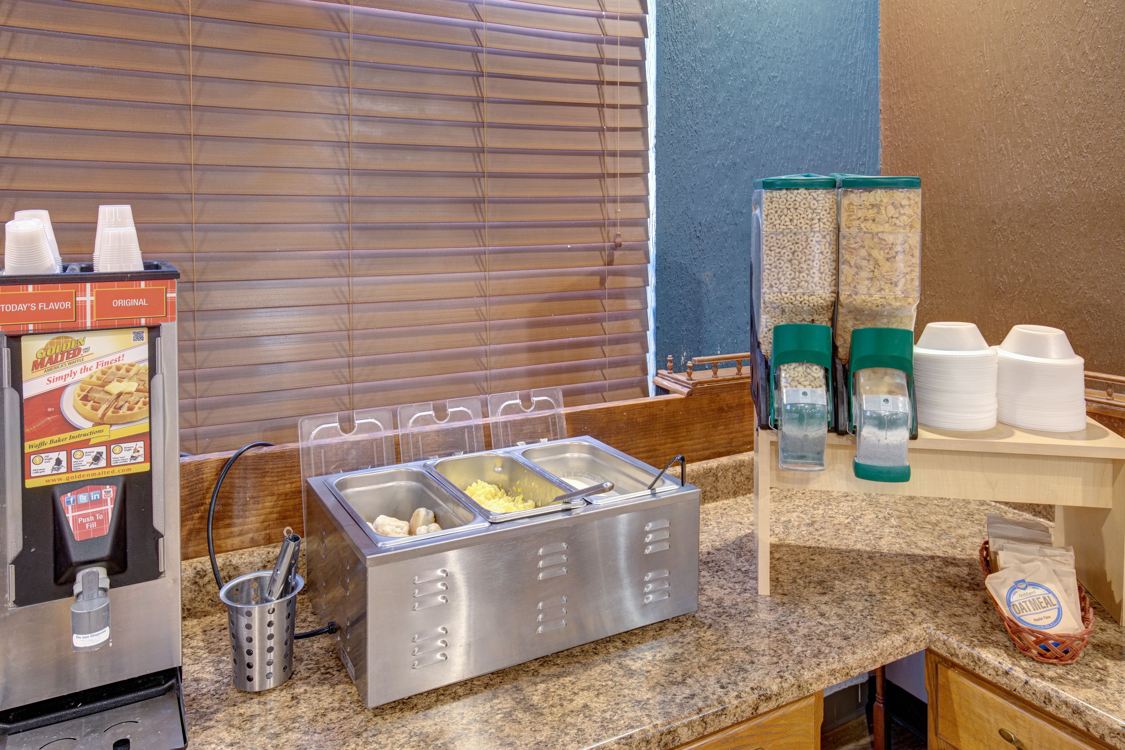 A self-service breakfast station with a waffle maker, beverage dispensers, and a coffee machine.