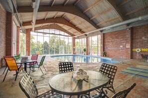 An indoor swimming pool area with a glass table and chairs, surrounded by brick walls and large windows.