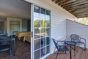 A room with a bed, a table, and two chairs. There is a sliding glass door leading to an outdoor area with a wooden deck.