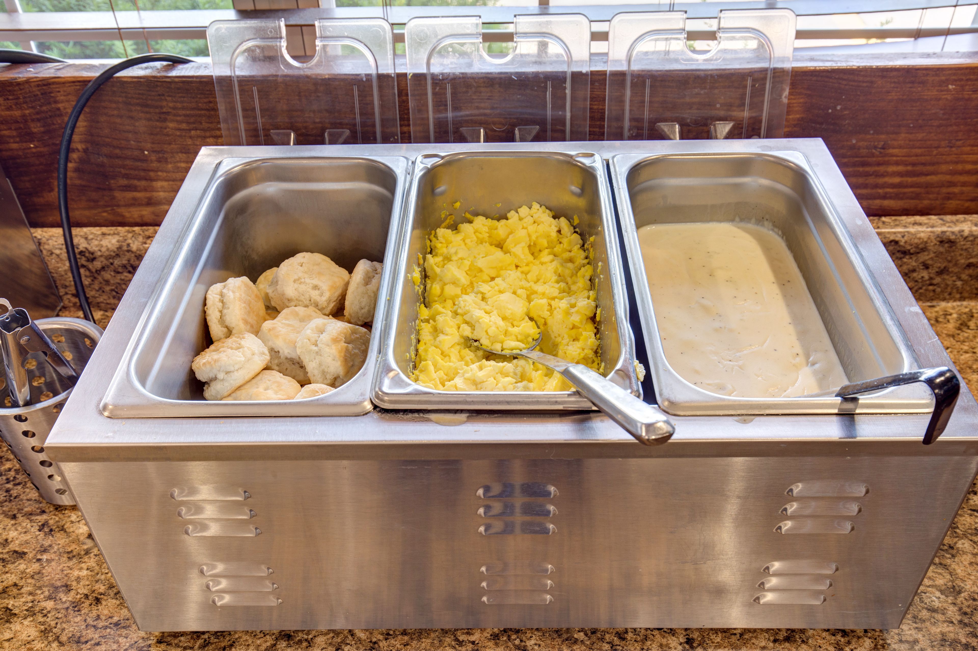 A buffet setup with three stainless steel trays containing biscuits, mashed potatoes, and gravy.