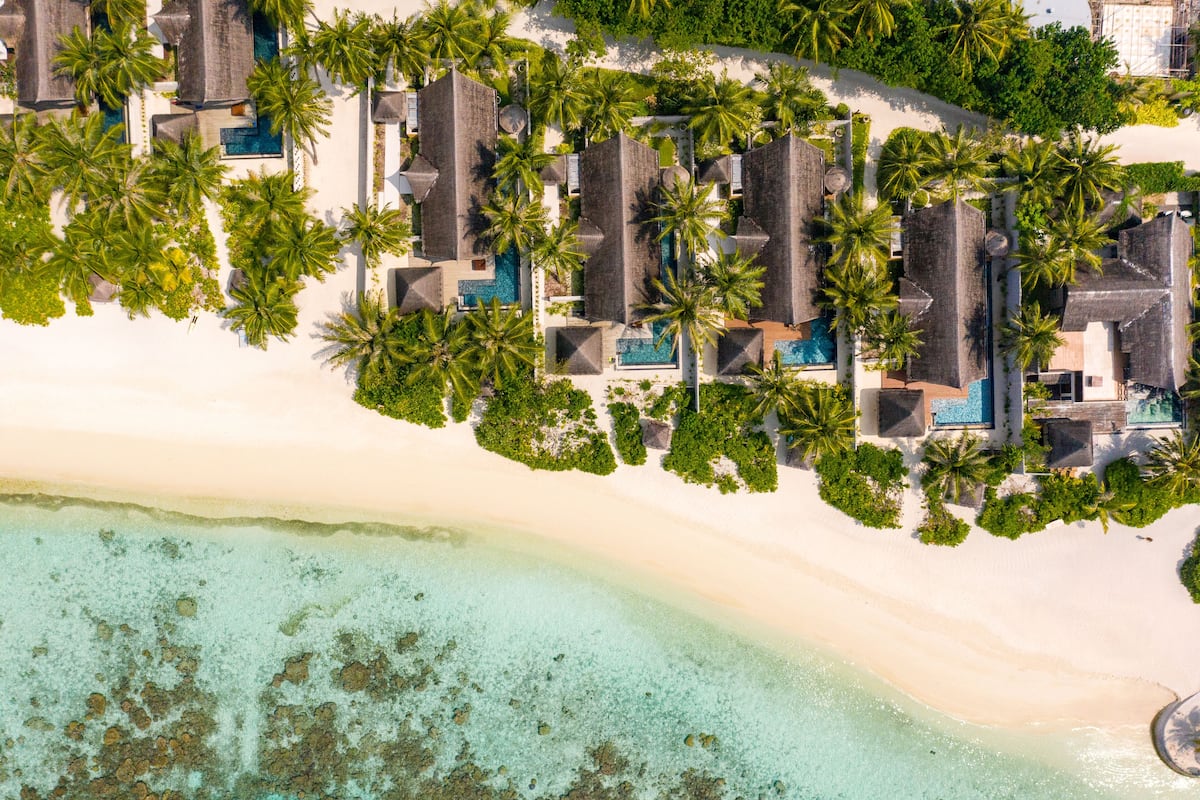 An aerial view of a resort with multiple overwater bungalows, a sandy beach, and clear turquoise waters.
