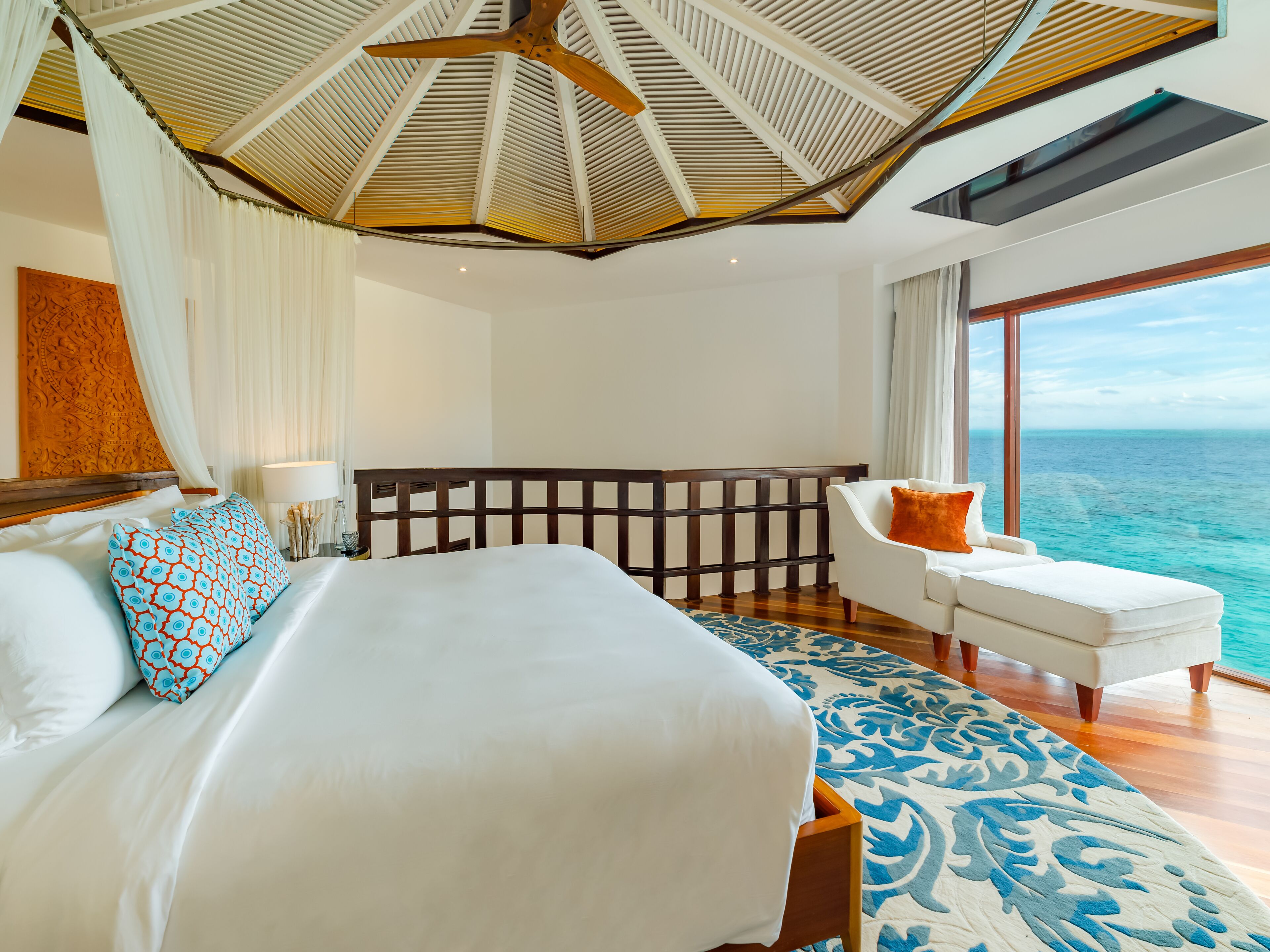 Habitación de hotel con una cama grande, cabecera de madera, un sillón blanco y vista al océano.