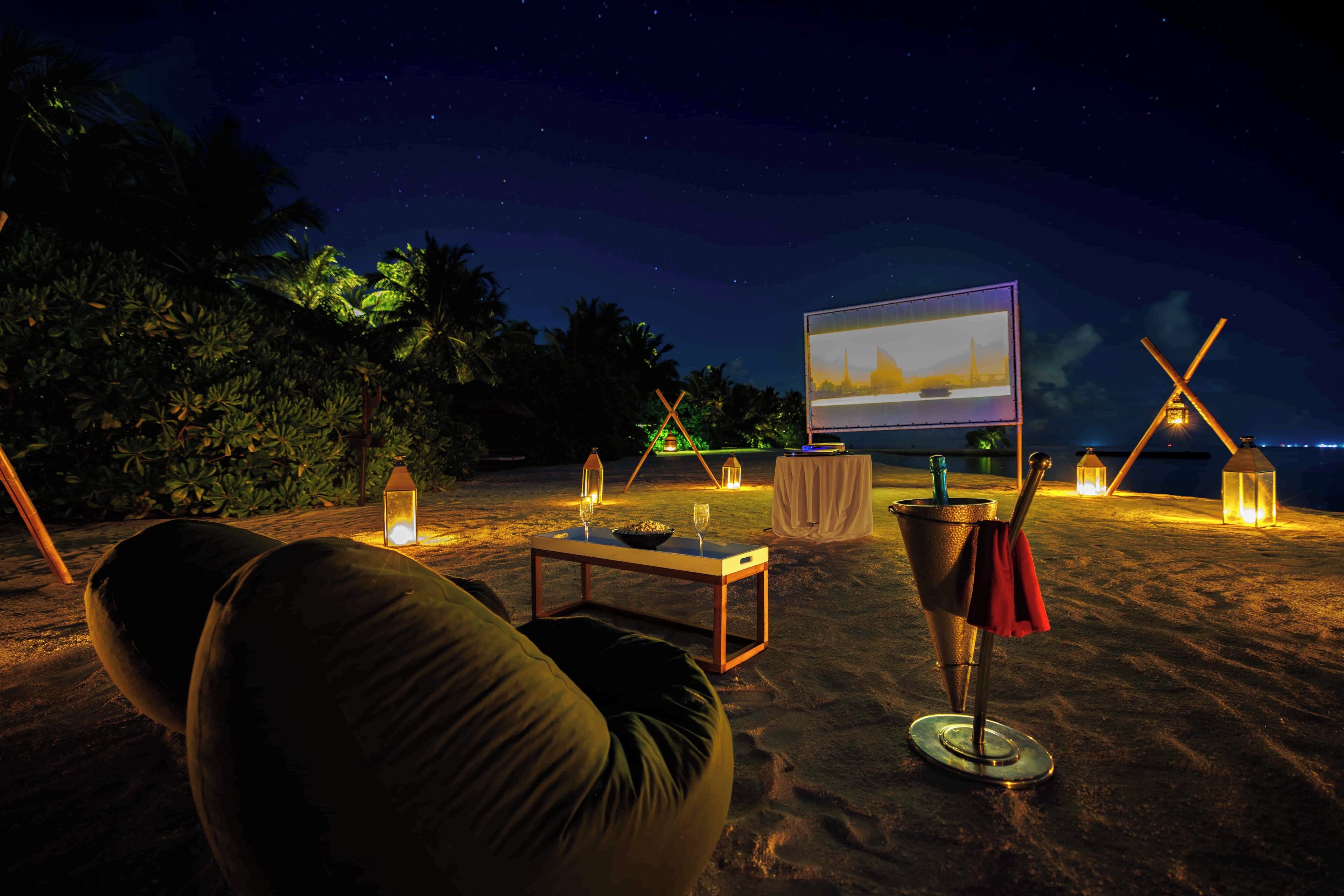 Escena de playa con una mesa, sillas y una pantalla proyectando un paisaje urbano.
