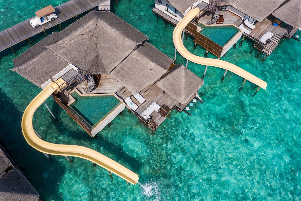 An aerial view of overwater bungalows with thatched roofs, clear turquoise water, and a yellow water slide.