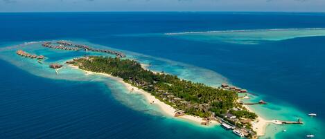 Vista aérea de una isla tropical con bungalows sobre el agua, aguas turquesas cristalinas y una pequeña zona de playa.