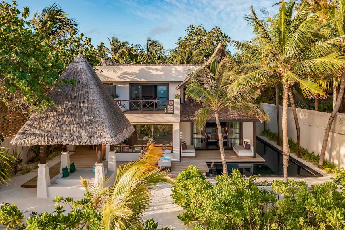 A two-story resort with a thatched-roof structure, a pool, and lush tropical vegetation.