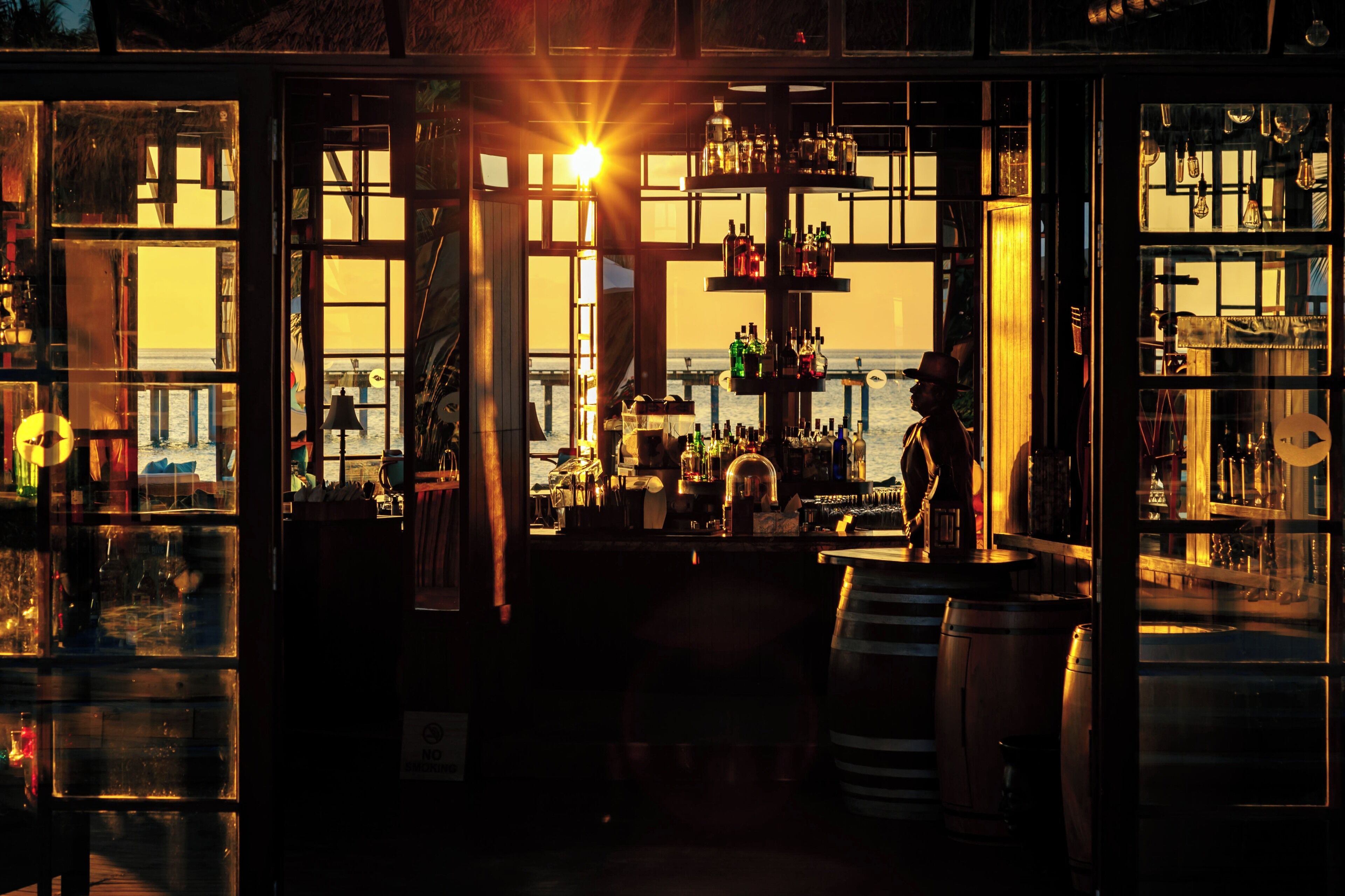 A bar with a view of the sea, shelves with bottles, and a person standing by the bar.