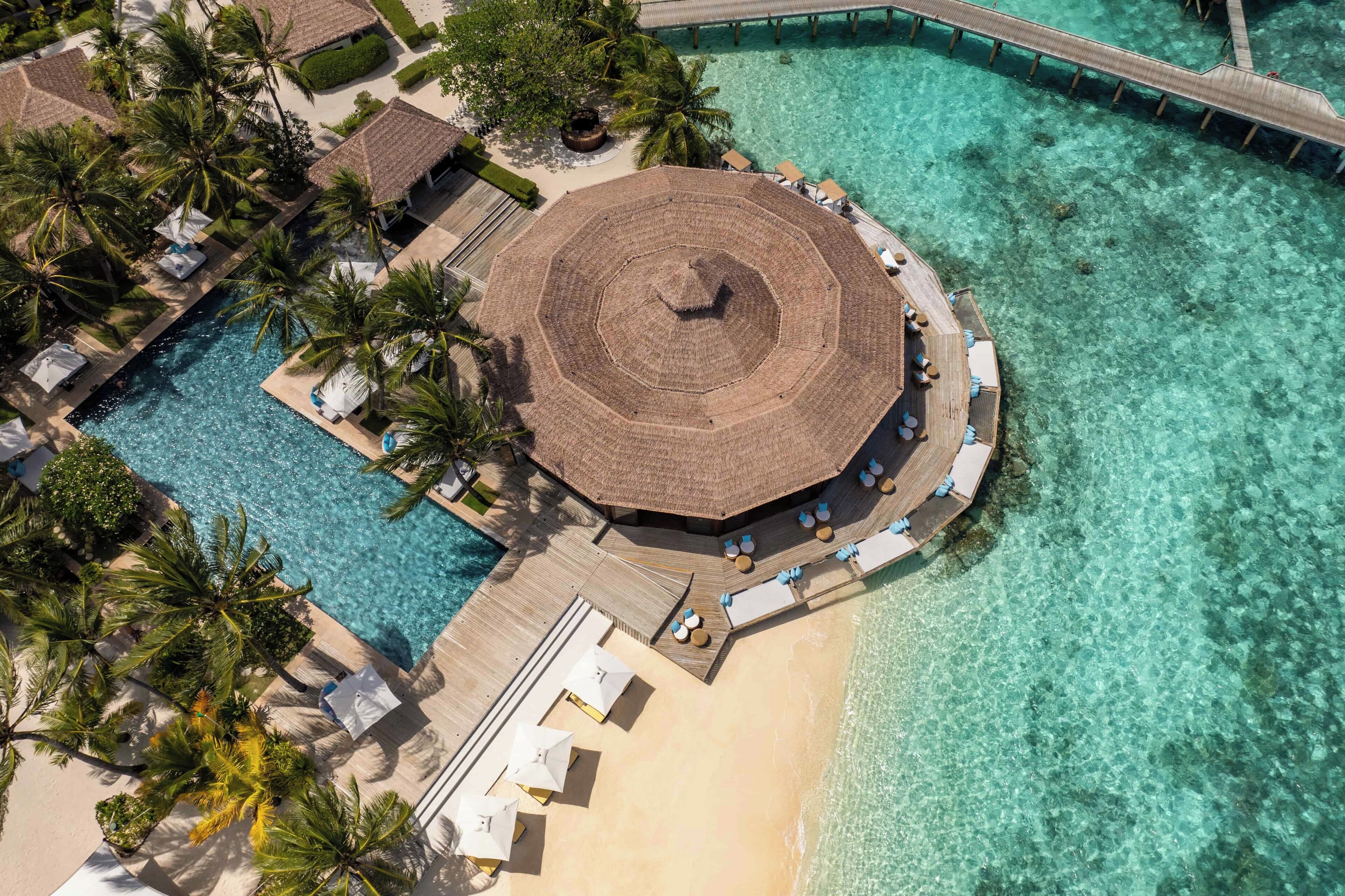 An aerial view of a resort with a circular building surrounded by a pool, a beach, and a pier.