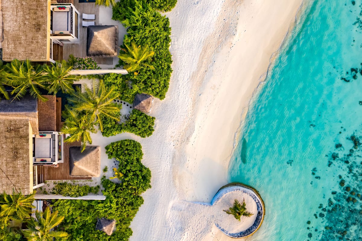 An aerial view of a beachside resort with thatched-roof huts, lush greenery, and clear turquoise waters.