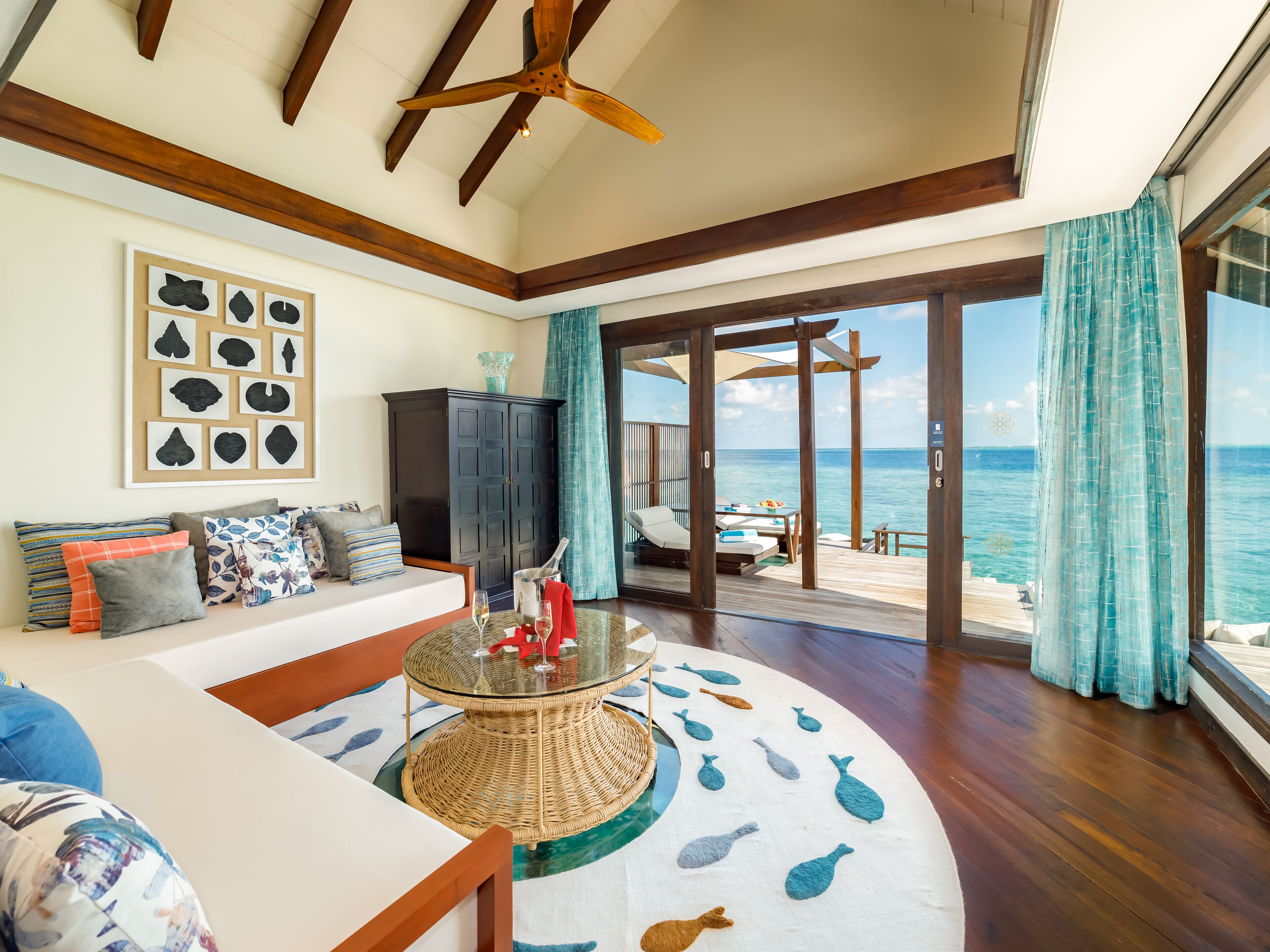 A spacious living area with a wooden ceiling fan, a large wall art piece, and a view of the ocean through a glass door.