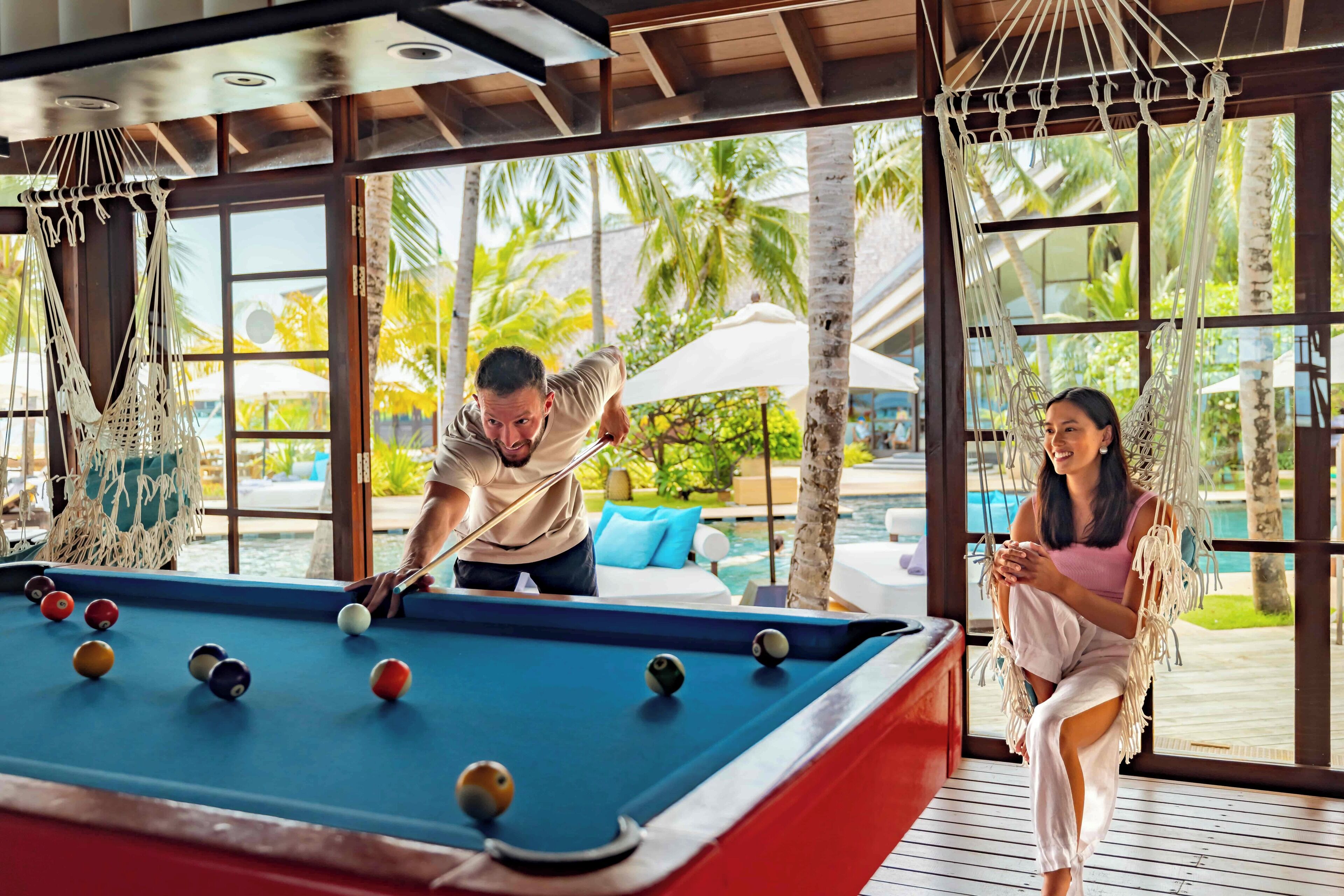 Uma mesa de bilhar em um quarto com grandes janelas, uma rede e vista para a piscina.