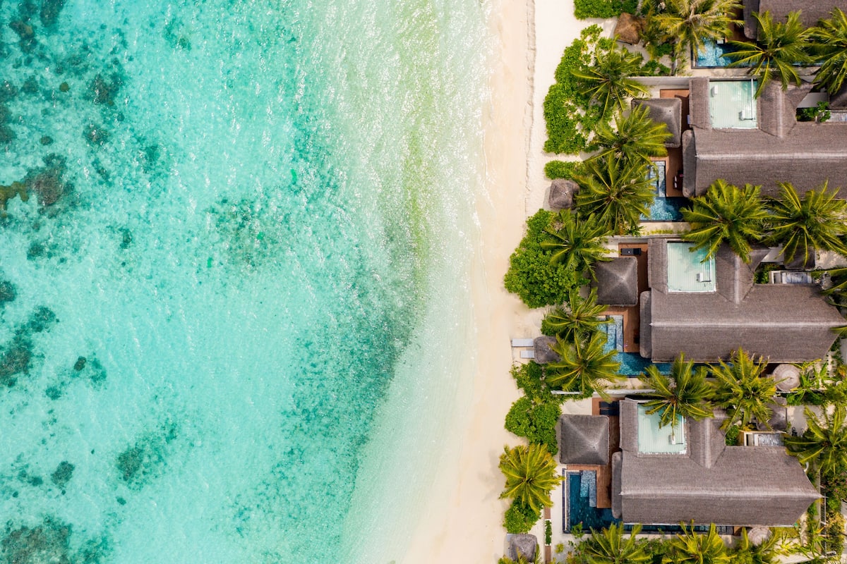 An aerial view of a beachfront resort with multiple overwater bungalows, clear turquoise waters, and lush greenery.