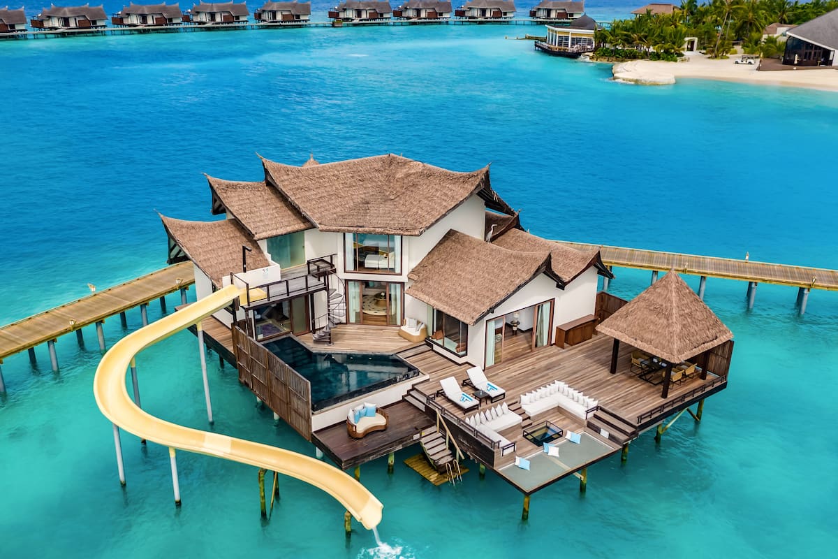 An overwater bungalow with a glass-enclosed infinity pool, wooden deck, and thatched-roof structures.