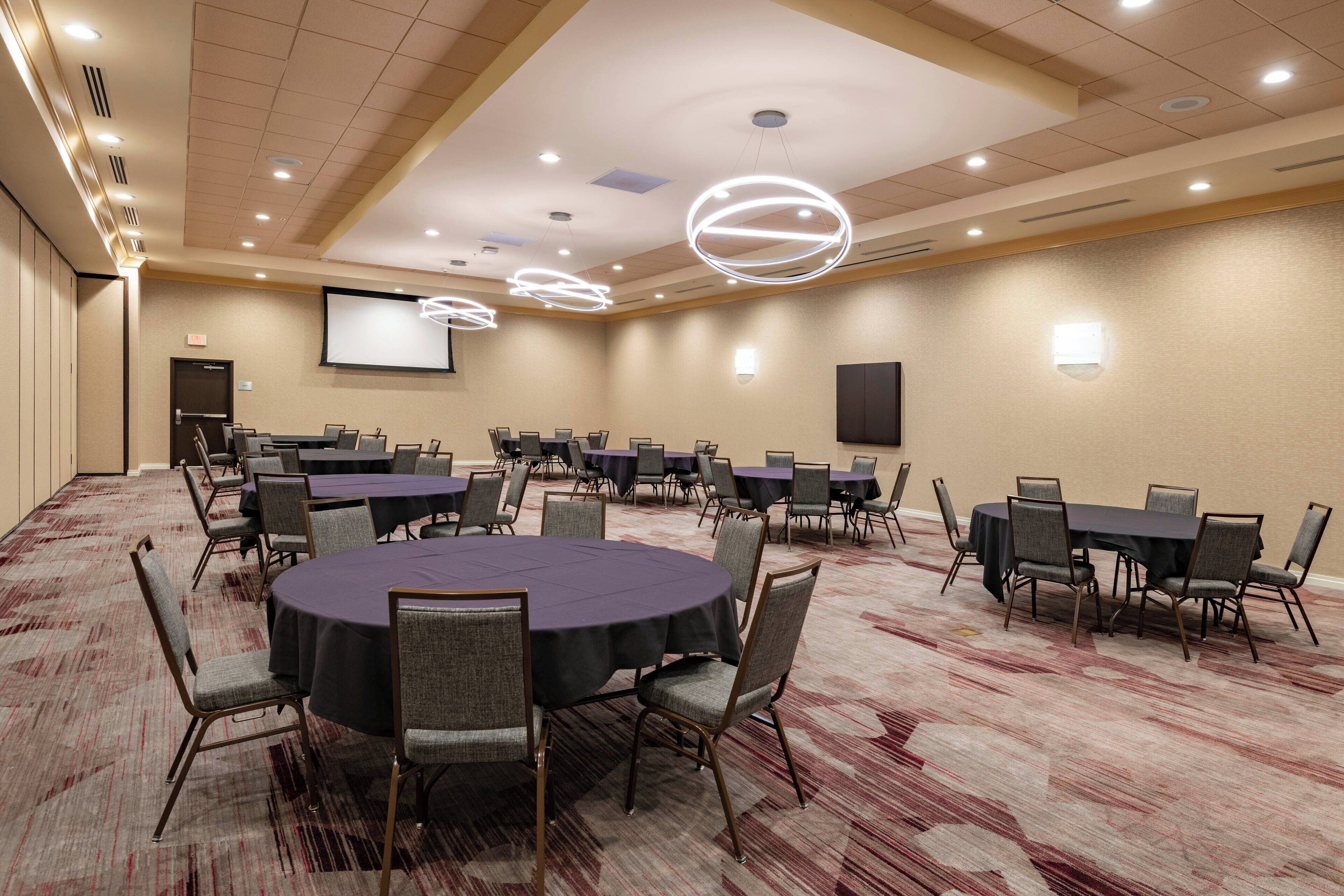 A conference room with multiple tables and chairs, a projector screen, and a carpeted floor.