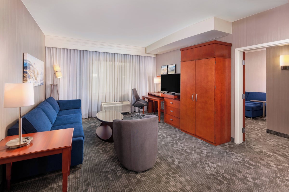 Habitación de hotel con un sofá azul, una mesita, un otoman gris, un armario de madera y un televisor.