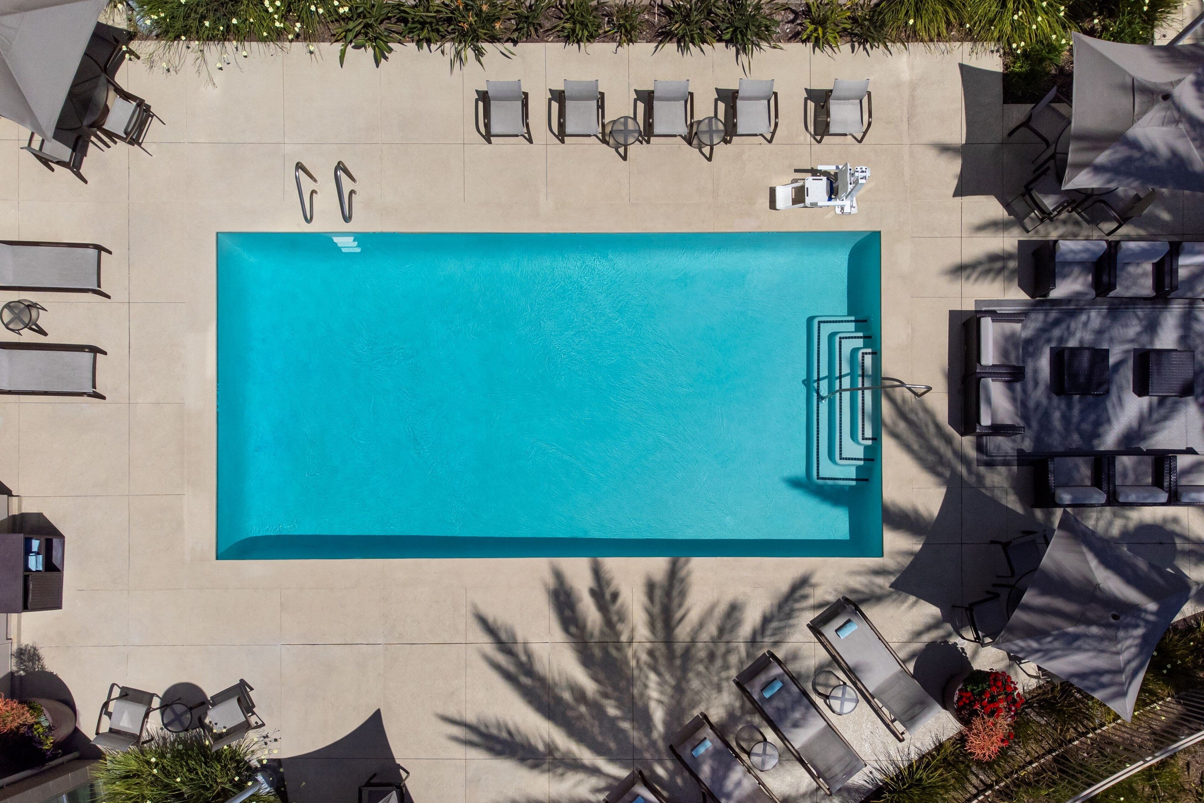 An aerial view of a rectangular swimming pool surrounded by lounge chairs and umbrellas, with a tiled deck and palm trees in the background.