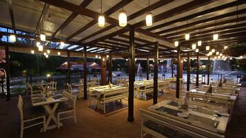 Un restaurante al aire libre con mesas y sillas blancas, lámparas colgantes y una vista a una fuente al fondo.
