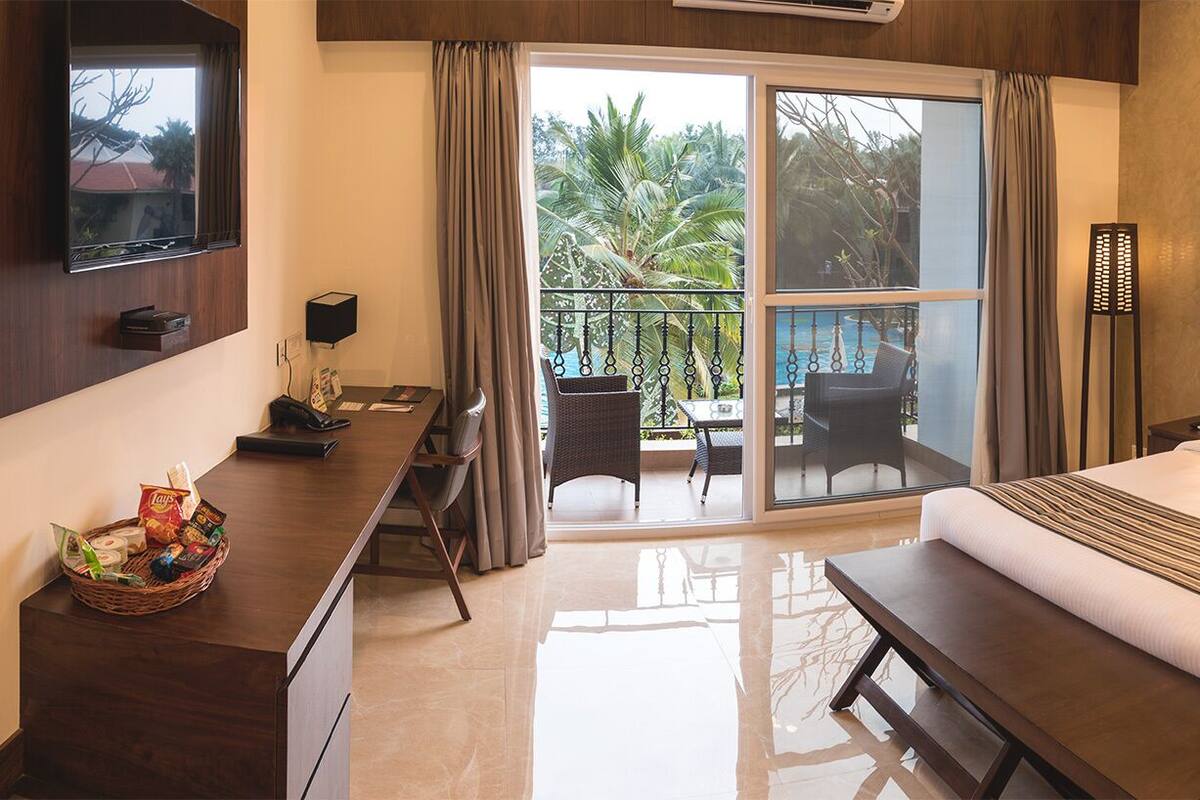 Habitación de hotel con una cama grande, un escritorio con una cesta de aperitivos, una silla y un balcón con vistas a palmeras y una piscina.