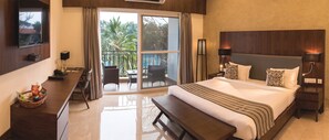 A hotel room with a large bed, a desk with a basket of snacks, a chair, and a balcony with a view of palm trees and a pool. - Silent Shores Resort & Spa (Mysore)