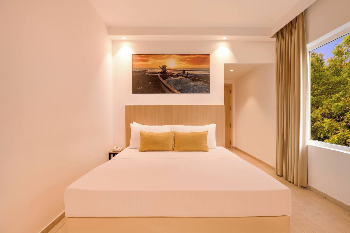 A hotel room with a large bed, a headboard, a nightstand, a painting of a sunset over the ocean, and a window with curtains.