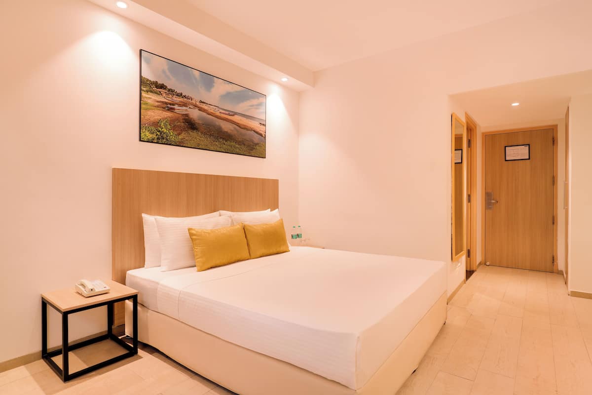 A modern hotel room with a large bed, a bedside table with a phone, a framed picture on the wall, and a wooden door.