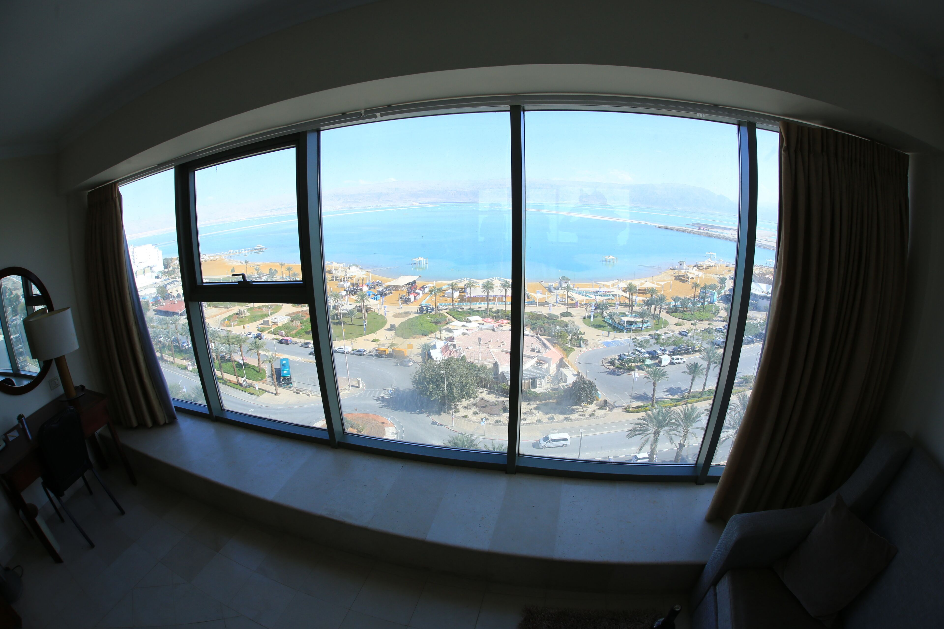 A room with a large curved window offering a view of a coastal area, including a beach and a road with vehicles.